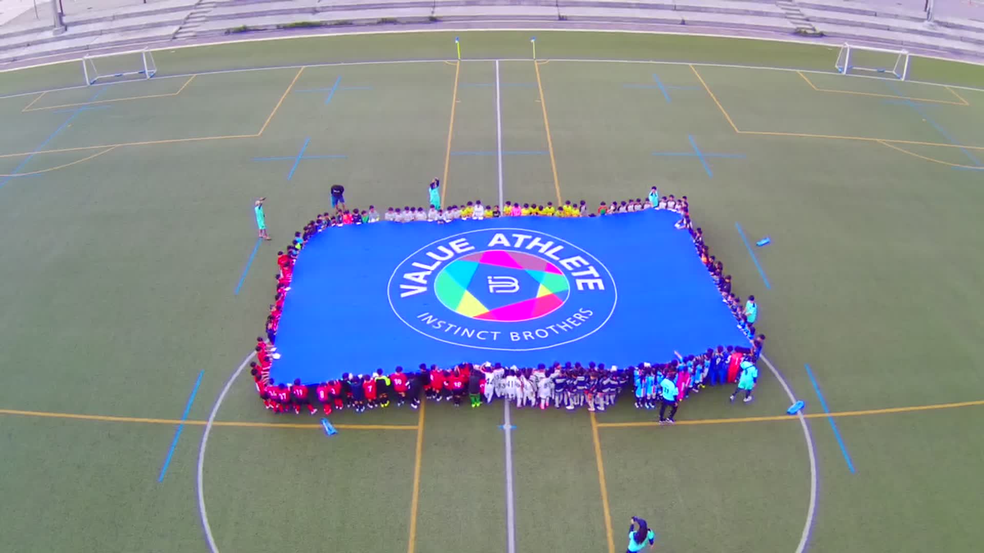 Hundreds of young athletes hold the VALUE ATHLETE flag together — the group's long-running youth sport and community programme.